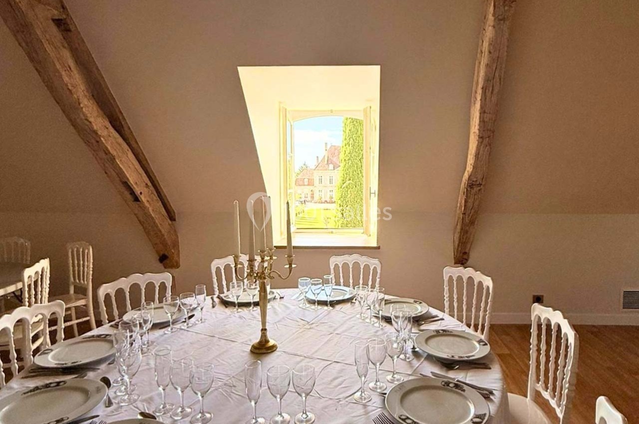 Table ronde élégamment dressée avec vaisselle et chandelier, située dans une salle lumineuse aux poutres apparentes.