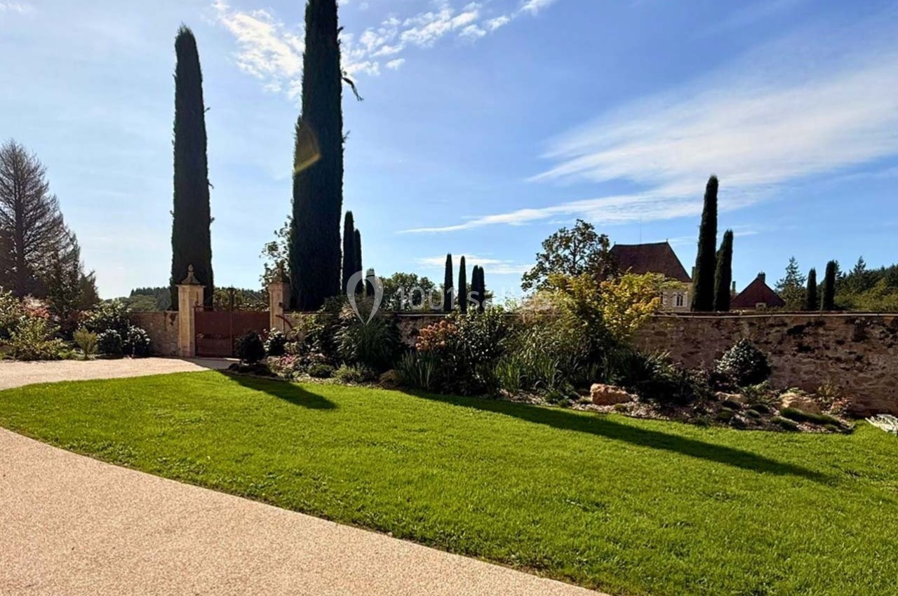 Allée en gravier bordée de pelouse et de cyprès, avec un mur en pierre et une maison en arrière-plan sous un ciel bleu.