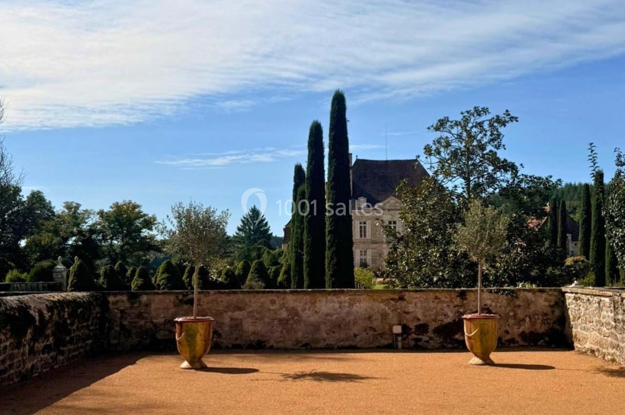 Terrasse en gravier avec deux pots d'arbres, vue sur un jardin arboré et un bâtiment ancien en arrière-plan.