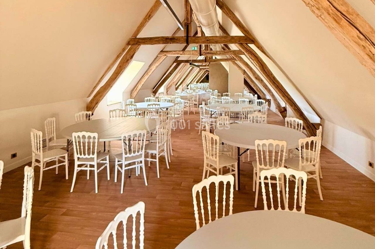 Salle mansardée avec poutres apparentes, tables rondes et chaises blanches disposées sur un sol en parquet clair.