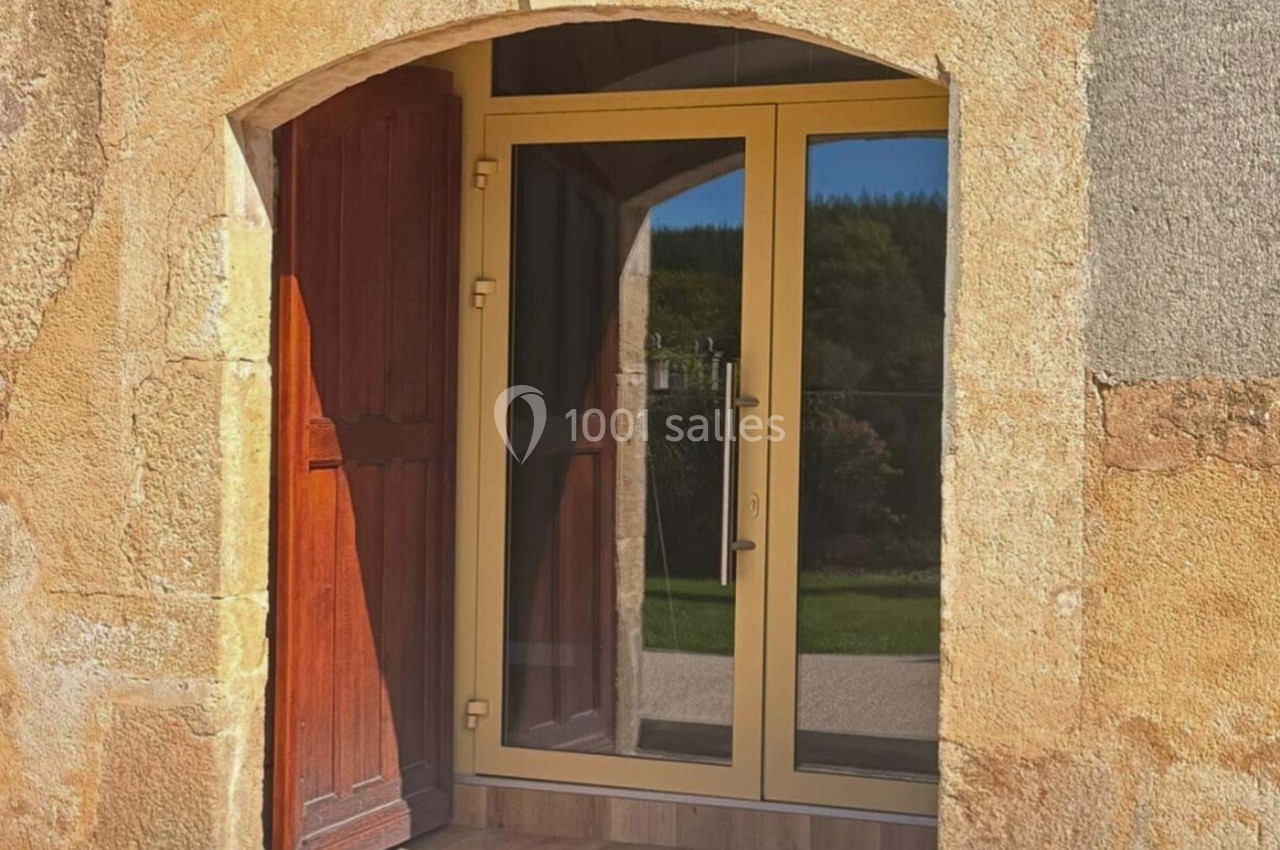 Porte vitrée moderne avec cadre doré, intégrée dans une façade en pierre ancienne, à côté d'une porte en bois ouverte.