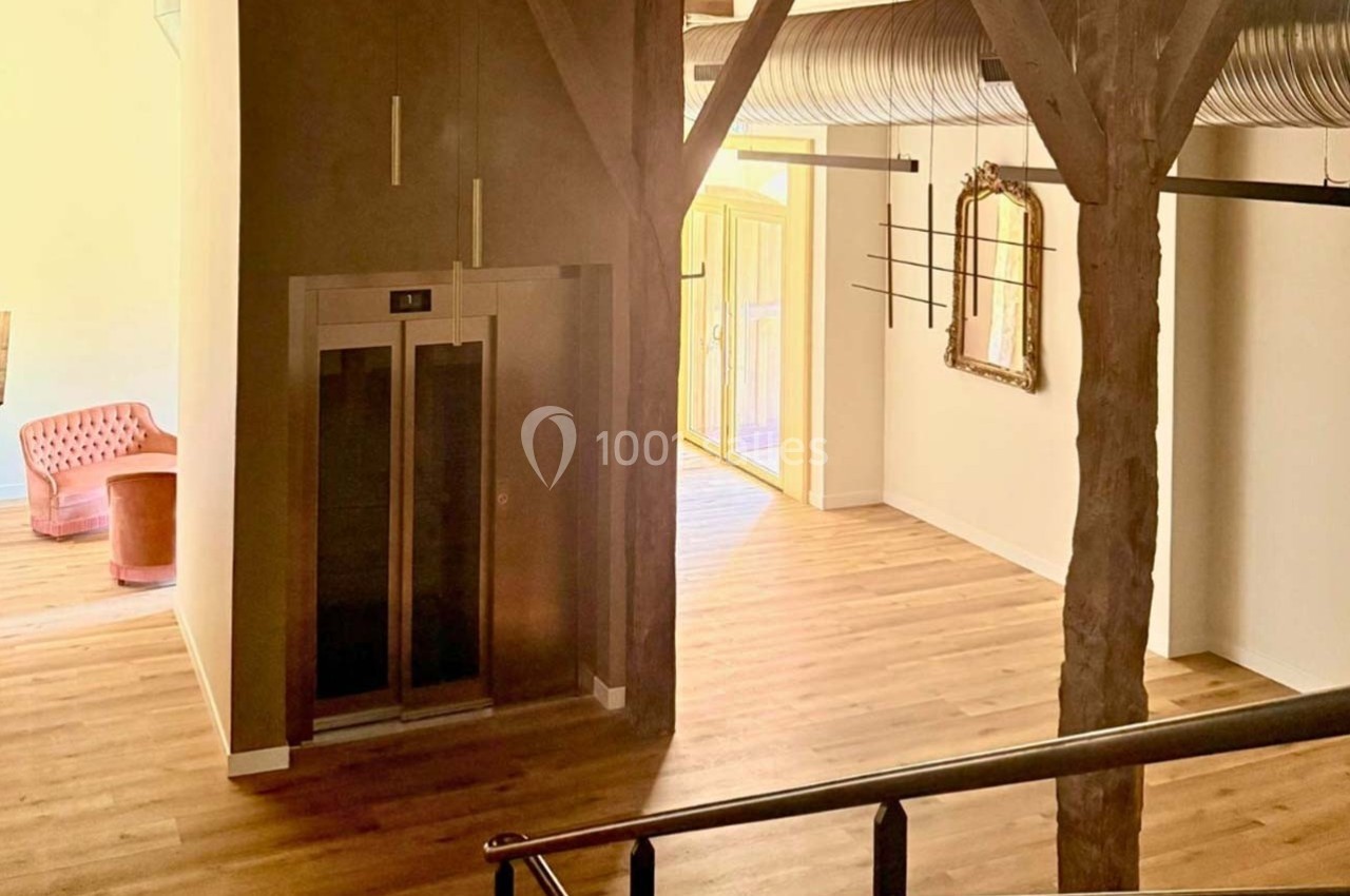 Intérieur lumineux avec parquet, poutres apparentes, ascenseur, fauteuil rouge et miroir sur un mur clair.