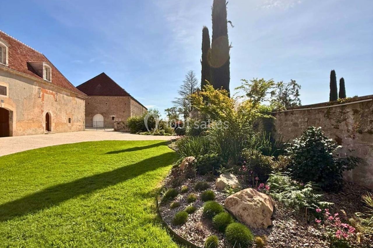 Jardin paysager avec pelouse, massifs de plantes et cyprès, bordé par des bâtiments en pierre sous un ciel ensoleillé.