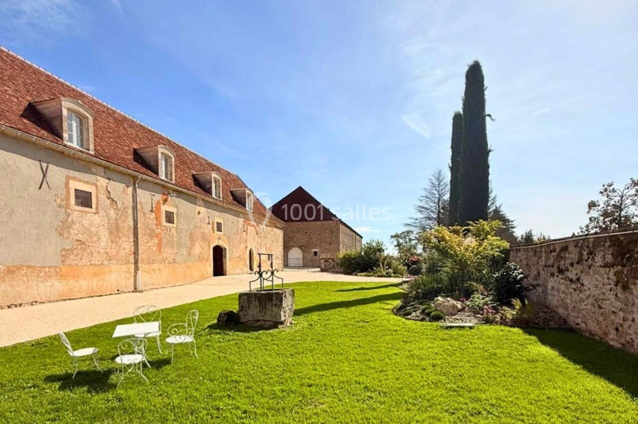 Cour verdoyante avec mobilier de jardin blanc, bordée de bâtiments anciens en pierre sous un ciel dégagé.