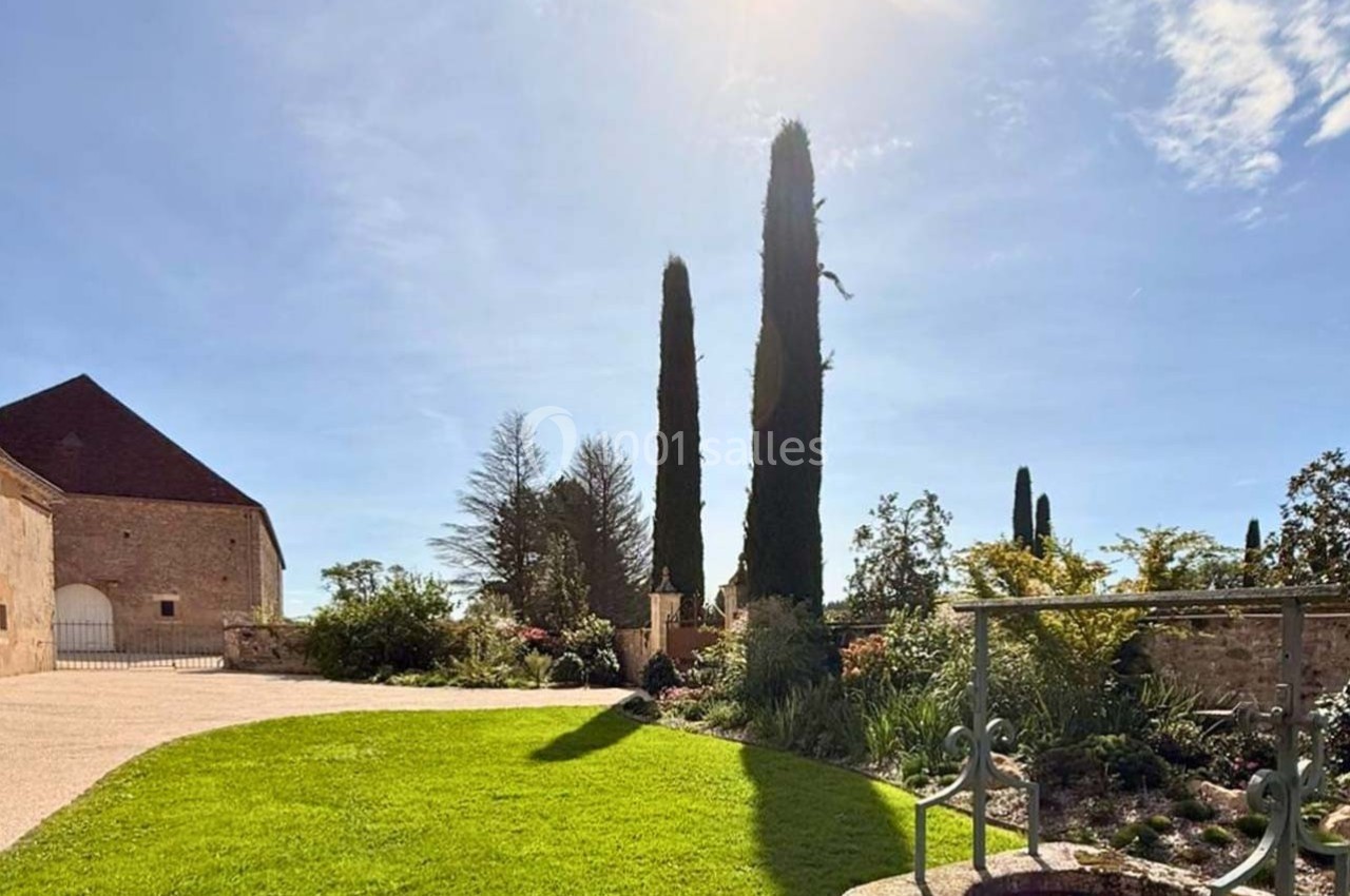 Jardin ensoleillé avec pelouse, puits en pierre, cyprès et bâtiment en pierre sous un ciel bleu dégagé.