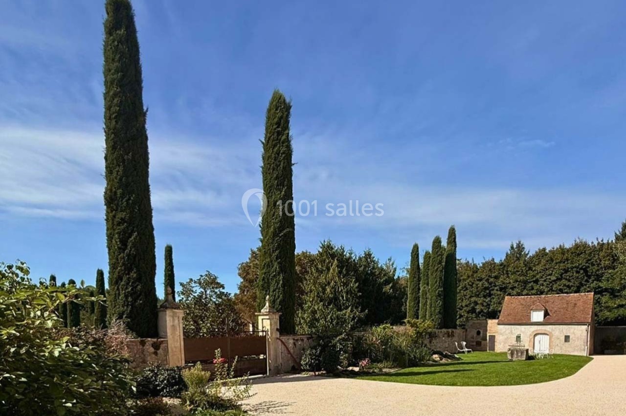 Allée bordée de cyprès menant à une maison en pierre avec un ciel bleu dégagé en arrière-plan.