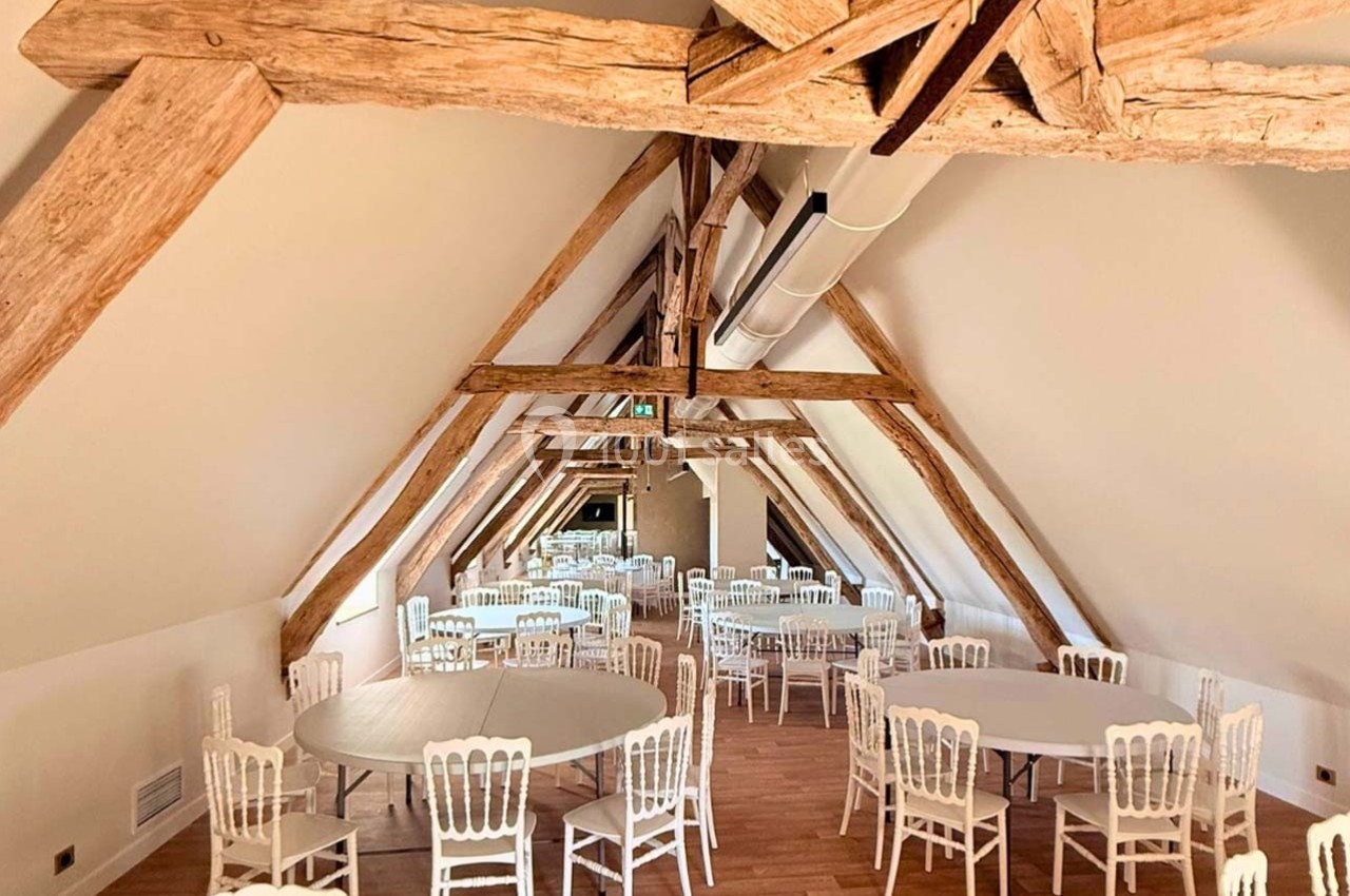 Salle mansardée avec poutres apparentes, tables rondes et chaises blanches disposées sur un sol en bois clair.