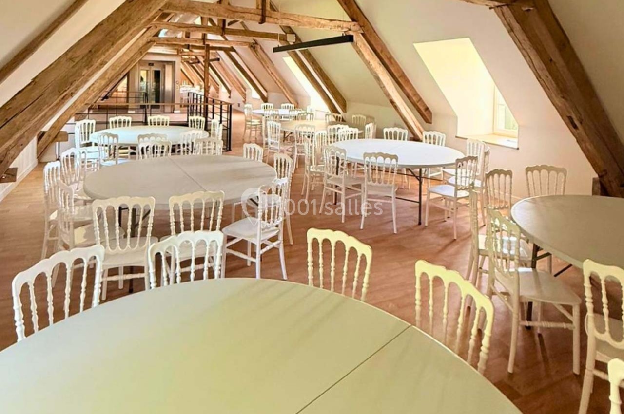 Salle lumineuse sous combles avec poutres apparentes, tables rondes blanches et chaises assorties disposées pour un…