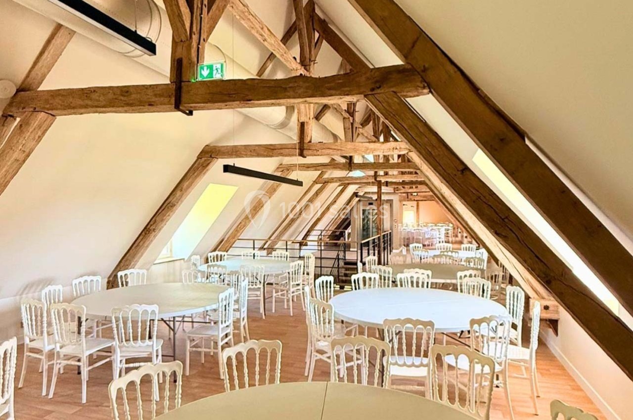 Salle lumineuse sous combles avec charpente en bois apparente, tables rondes et chaises blanches disposées pour un événement.