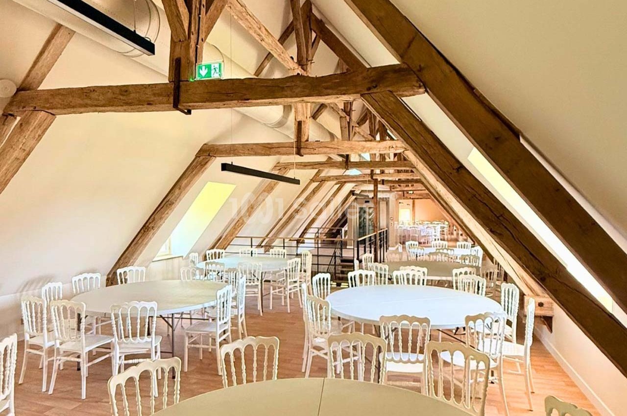 Salle lumineuse sous combles avec charpente en bois apparente, tables rondes et chaises blanches disposées pour un événement.