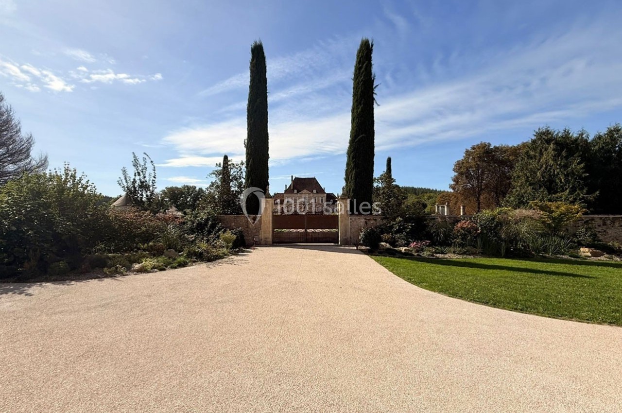 Allée en gravier menant à une maison entourée de jardins, avec deux grands cyprès encadrant l'entrée.