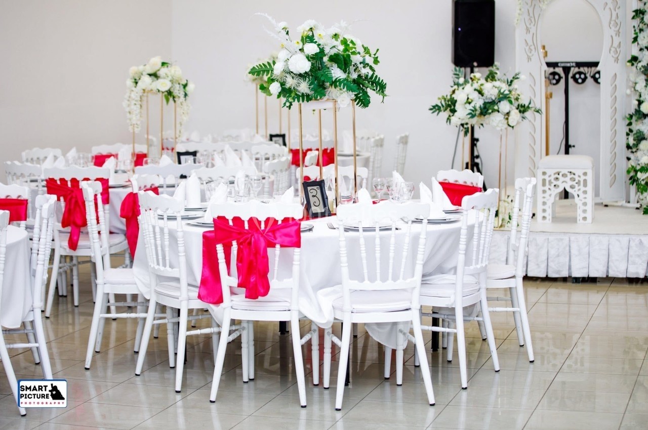 Salle de réception décorée avec des tables rondes, nappes blanches, chaises blanches ornées de nœuds rouges et arrangements…