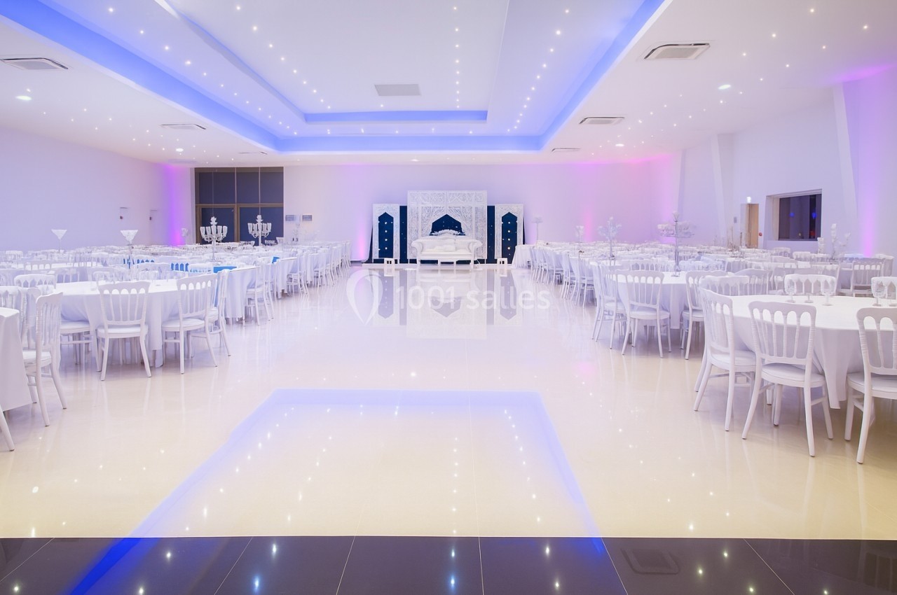 Salle de réception spacieuse avec tables rondes dressées, éclairage violet et blanc, et une estrade décorée.