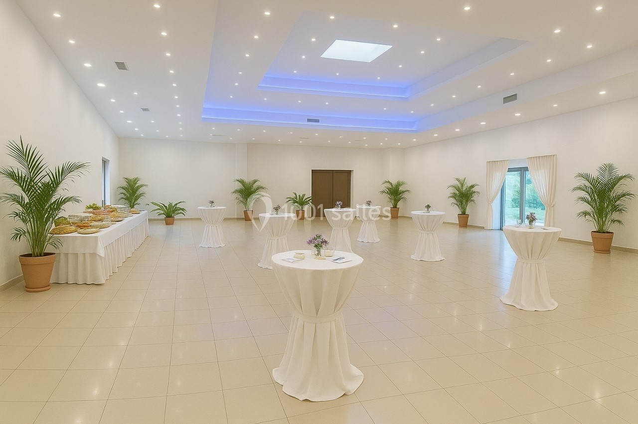 Salle lumineuse avec tables hautes drapées de blanc, plantes en pot et buffet disposé sur le côté.