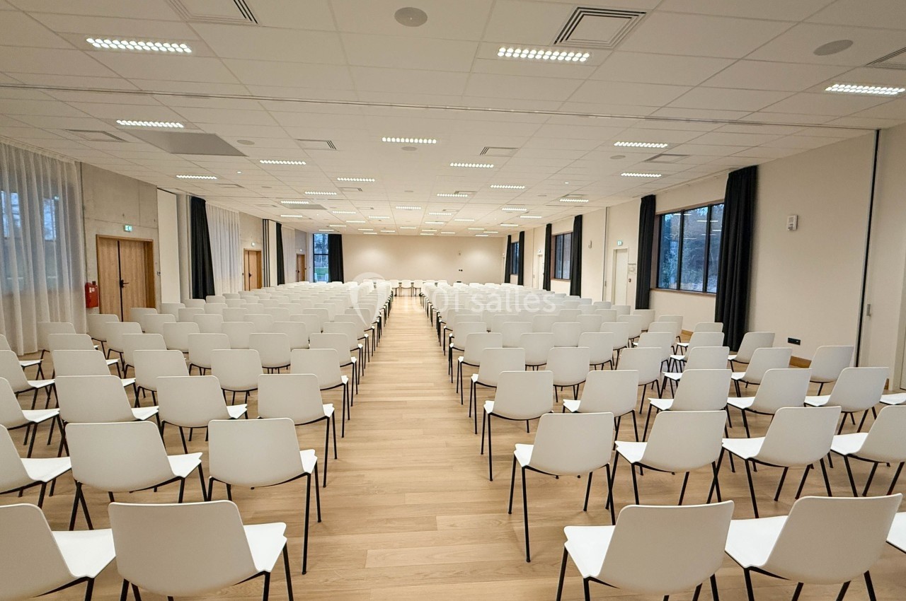 Salle de conférence vide avec rangées de chaises blanches alignées sur un sol en bois clair, éclairée par des plafonniers.
