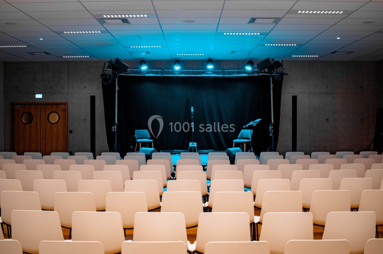 Salle de spectacle vide avec des rangées de chaises blanches face à une scène éclairée par des projecteurs bleus.