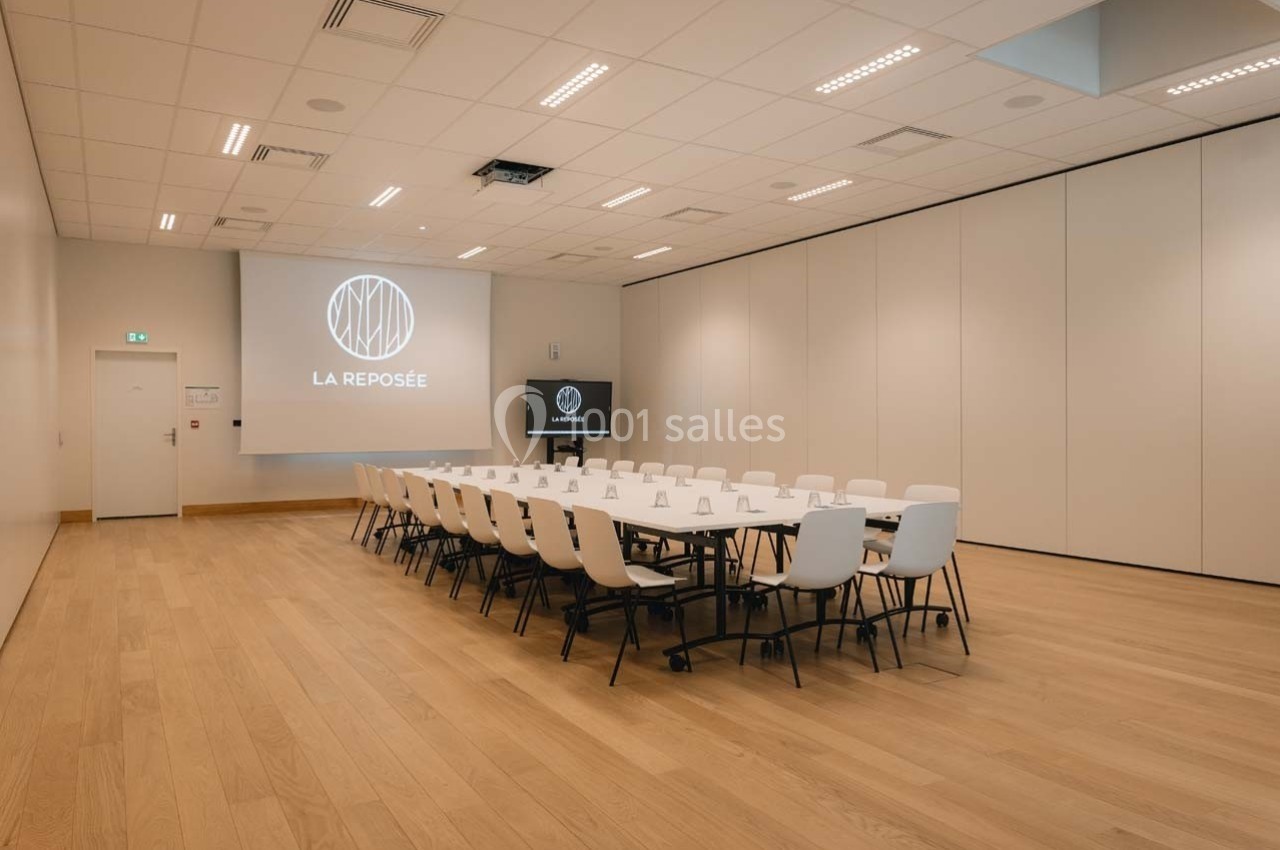 Salle de réunion moderne avec tables disposées en rectangle, chaises blanches, écran de projection et parquet clair.