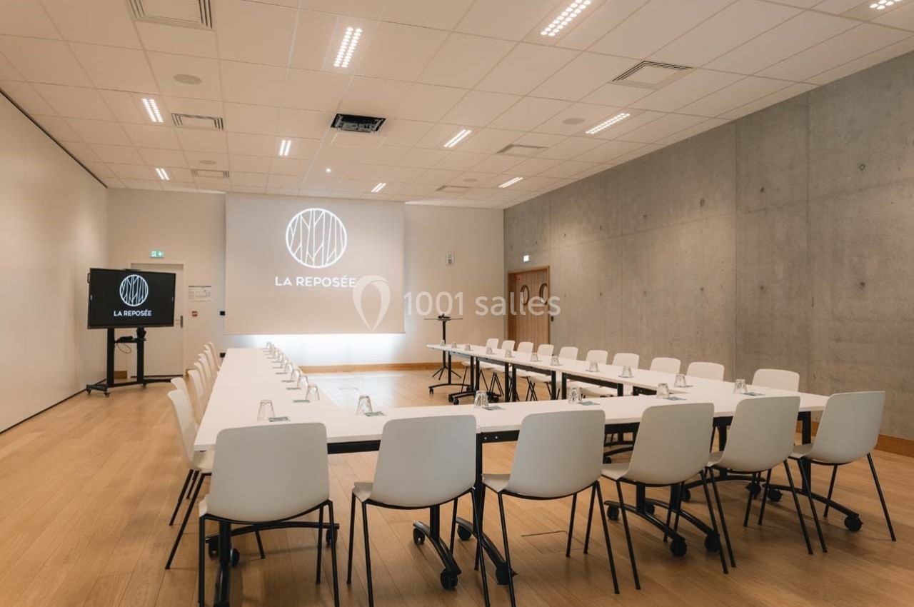 Salle de réunion lumineuse avec tables disposées en U, chaises blanches et écran de projection affichant ’La Reposée’.