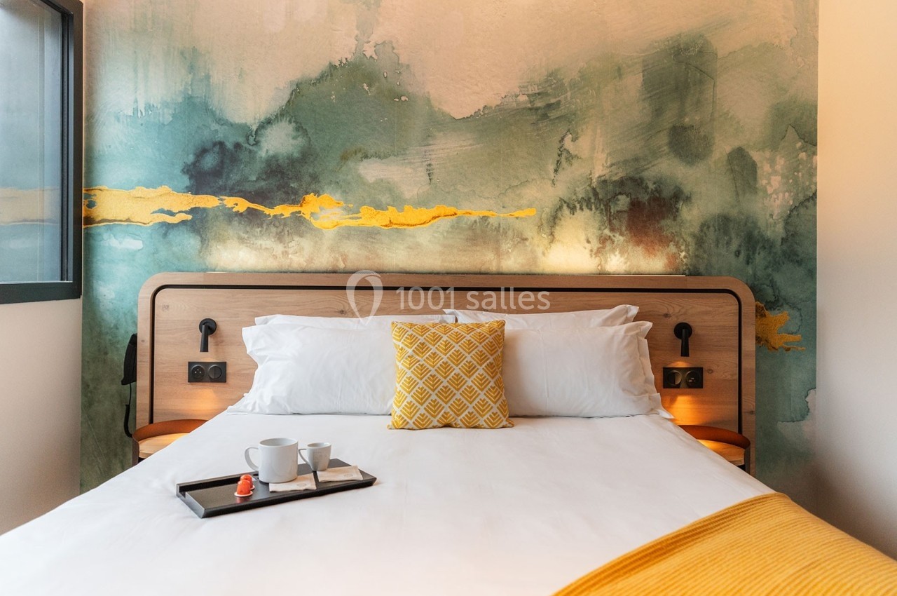 Lit double avec coussin jaune, plaid assorti, plateau avec tasses et fond de mur peint dans des tons verts et jaunes.