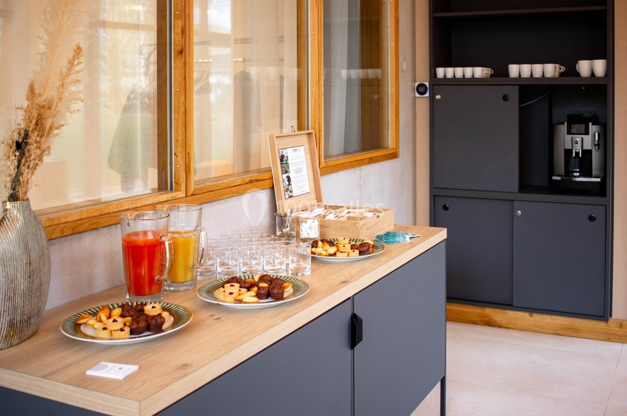 Buffet avec jus de fruits, biscuits et verres sur un meuble, près d'une machine à café et d'armoires grises.