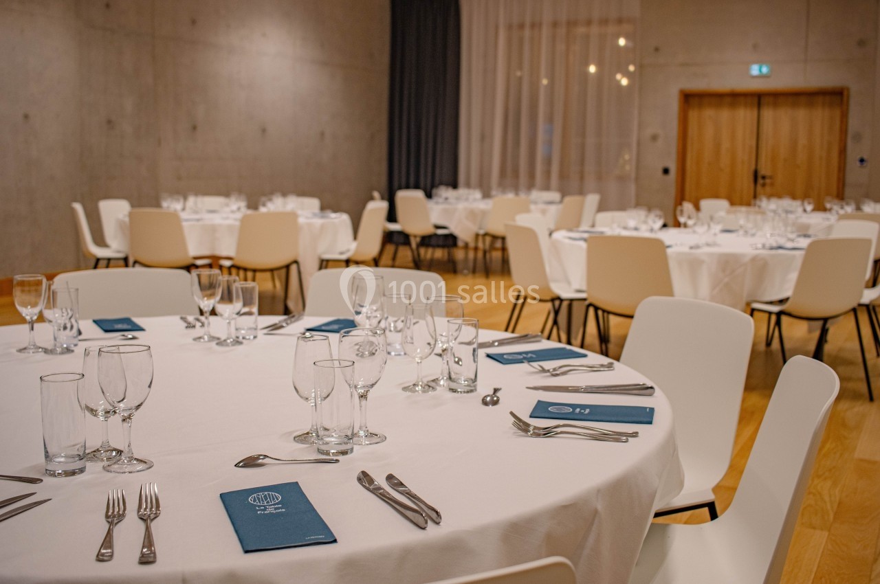 Salle de réception avec tables rondes dressées, nappes blanches, couverts, verres et menus bleus.