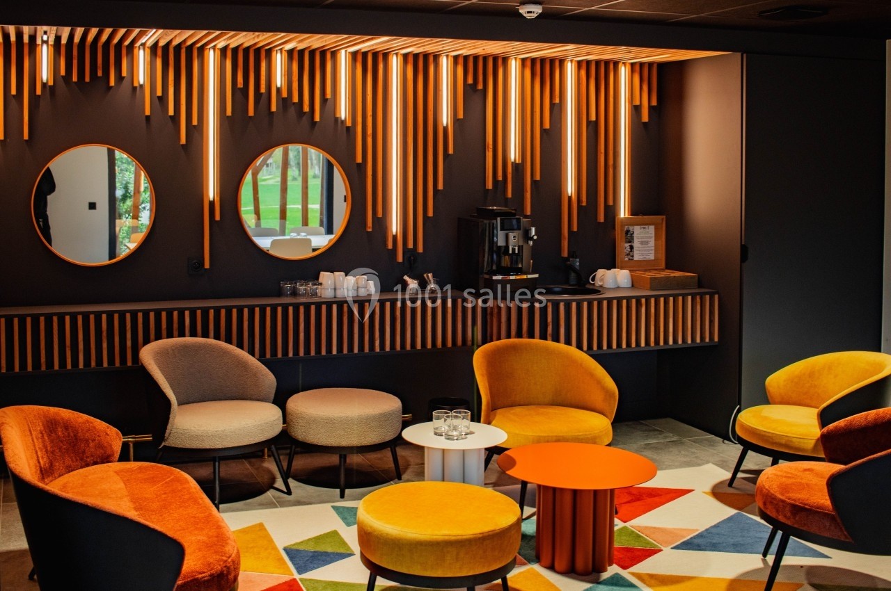 Espace lounge moderne avec fauteuils colorés, tables basses, machine à café et éclairage suspendu orange.