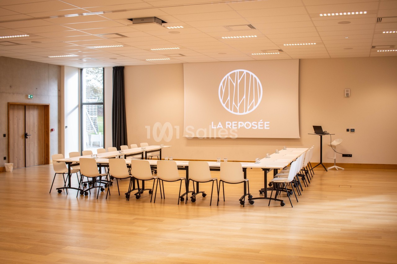 Salle de réunion lumineuse avec tables disposées en U, chaises blanches et écran de projection affichant ’La Reposée’.
