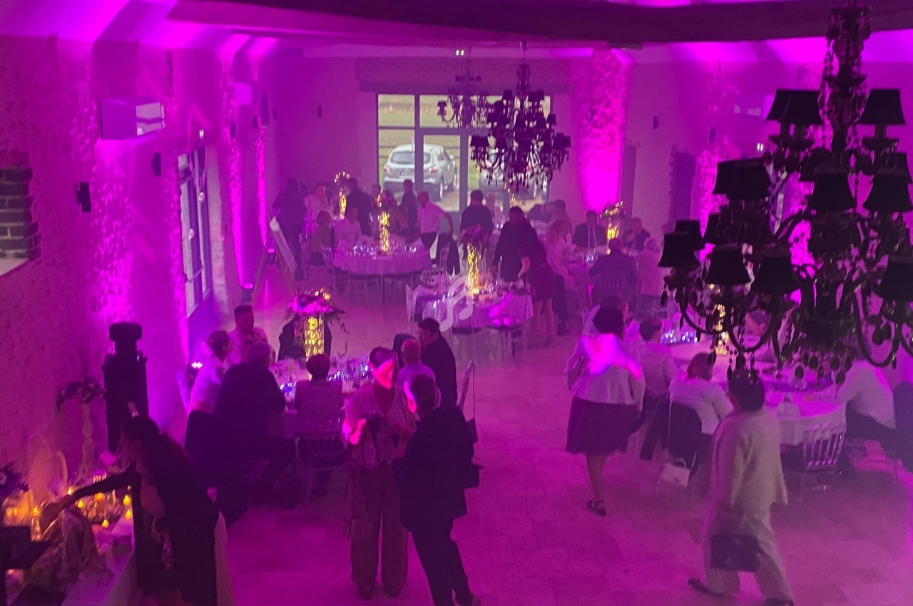 Salle de réception éclairée en rose avec des invités assis et debout autour de tables décorées.