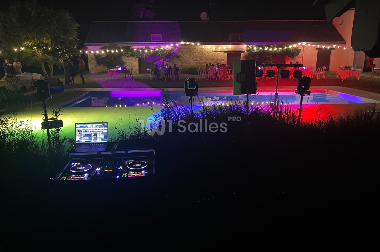 Console de DJ en premier plan face à une piscine éclairée de lumières colorées lors d'une soirée en extérieur.