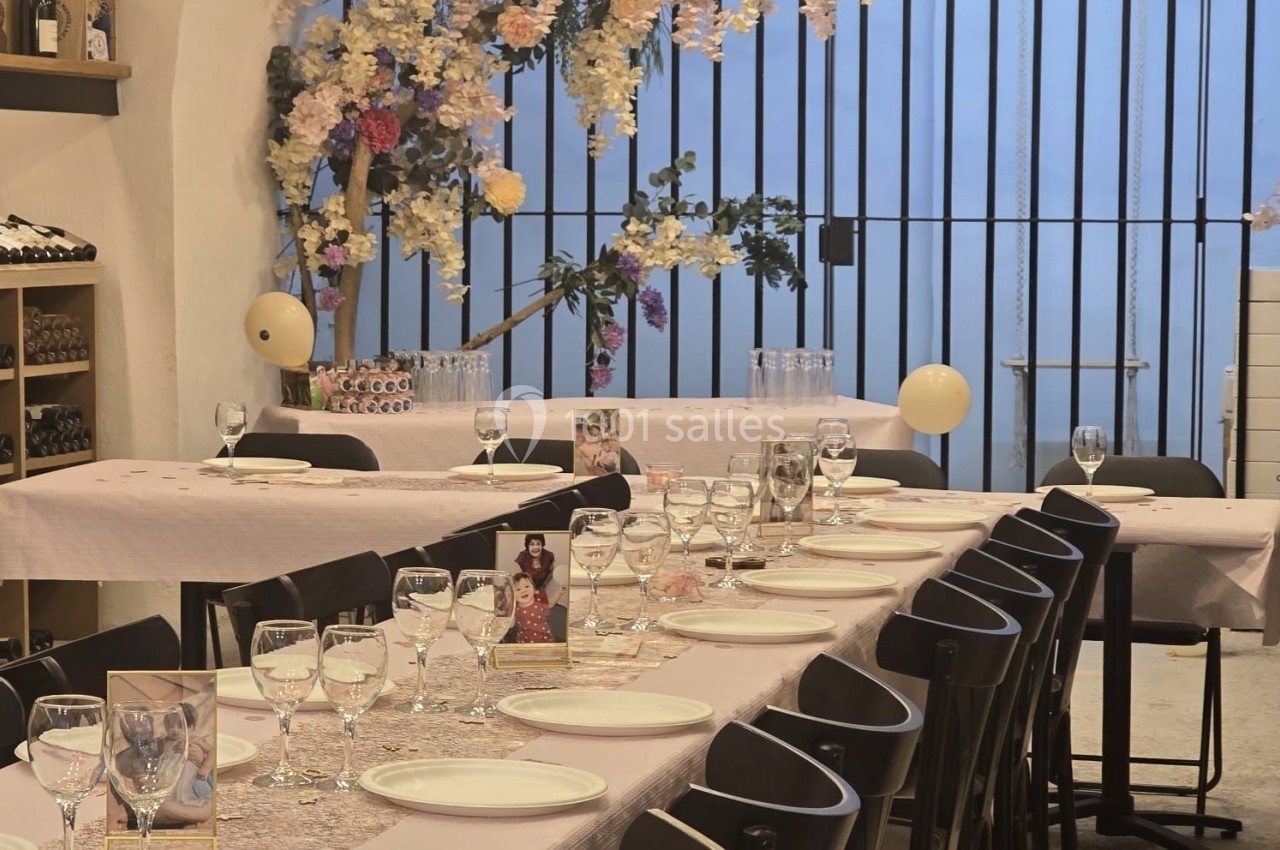 Salle décorée pour un événement avec tables dressées, nappes roses, vaisselle, verres et fleurs suspendues.