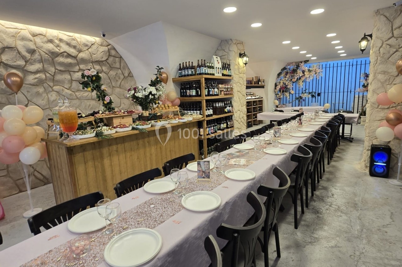 Salle décorée pour un événement, avec longues tables dressées, ballons, buffet et étagères de bouteilles en arrière-plan.