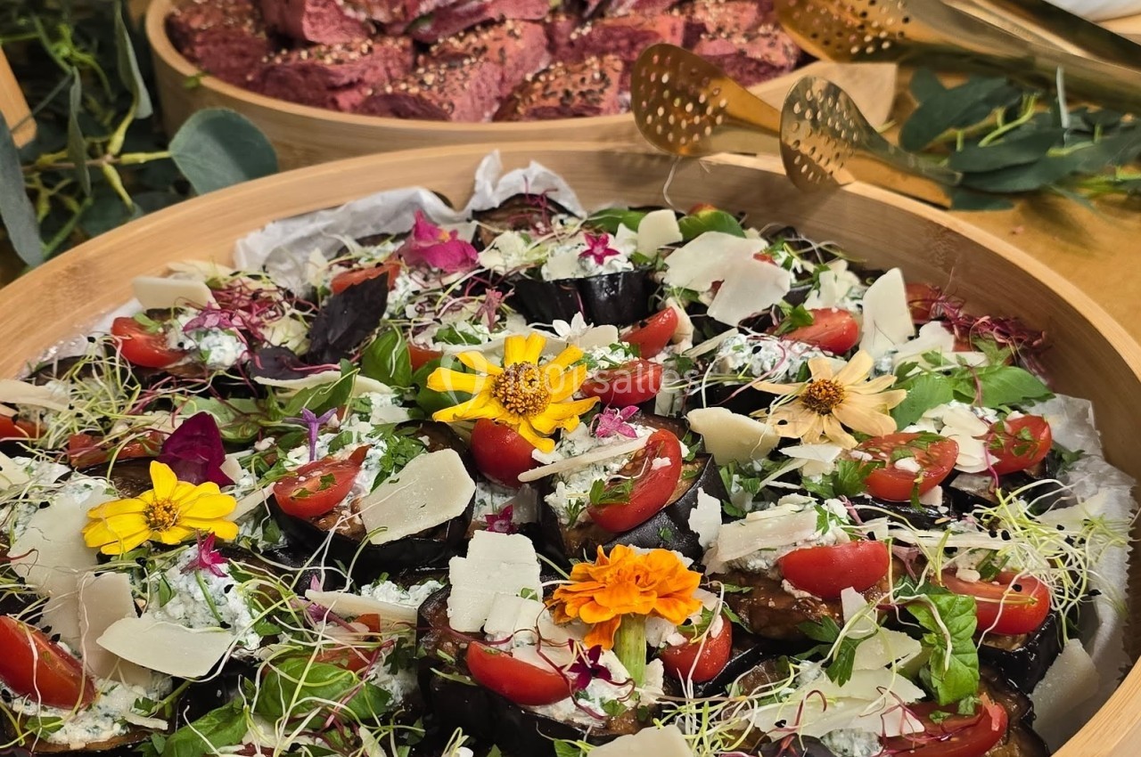 Plateau de légumes garnis de fromage, tomates cerises et fleurs comestibles, avec des pains colorés en arrière-plan.