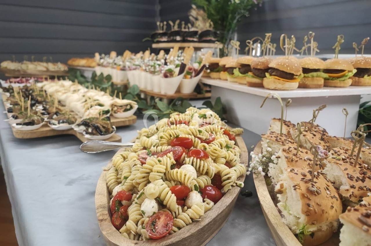 Buffet varié avec salade de pâtes, mini-burgers, pains garnis et autres plats disposés sur une table décorée.