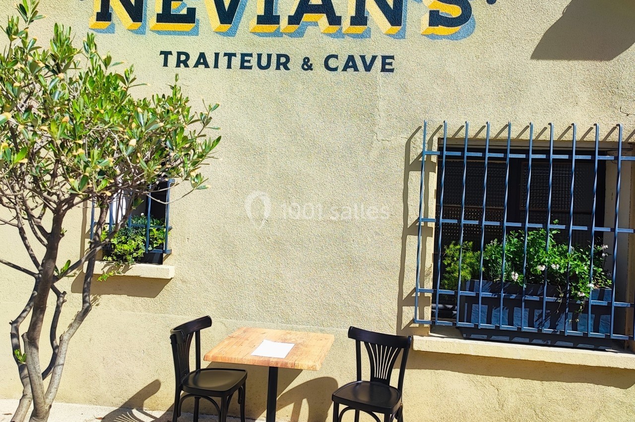 Table et deux chaises noires installées devant un mur beige avec une enseigne ’Nevian's Traiteur & Cave’.