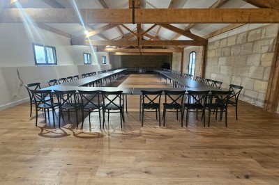 Salle spacieuse avec parquet en bois, poutres apparentes et tables disposées en U entourées de chaises noires.