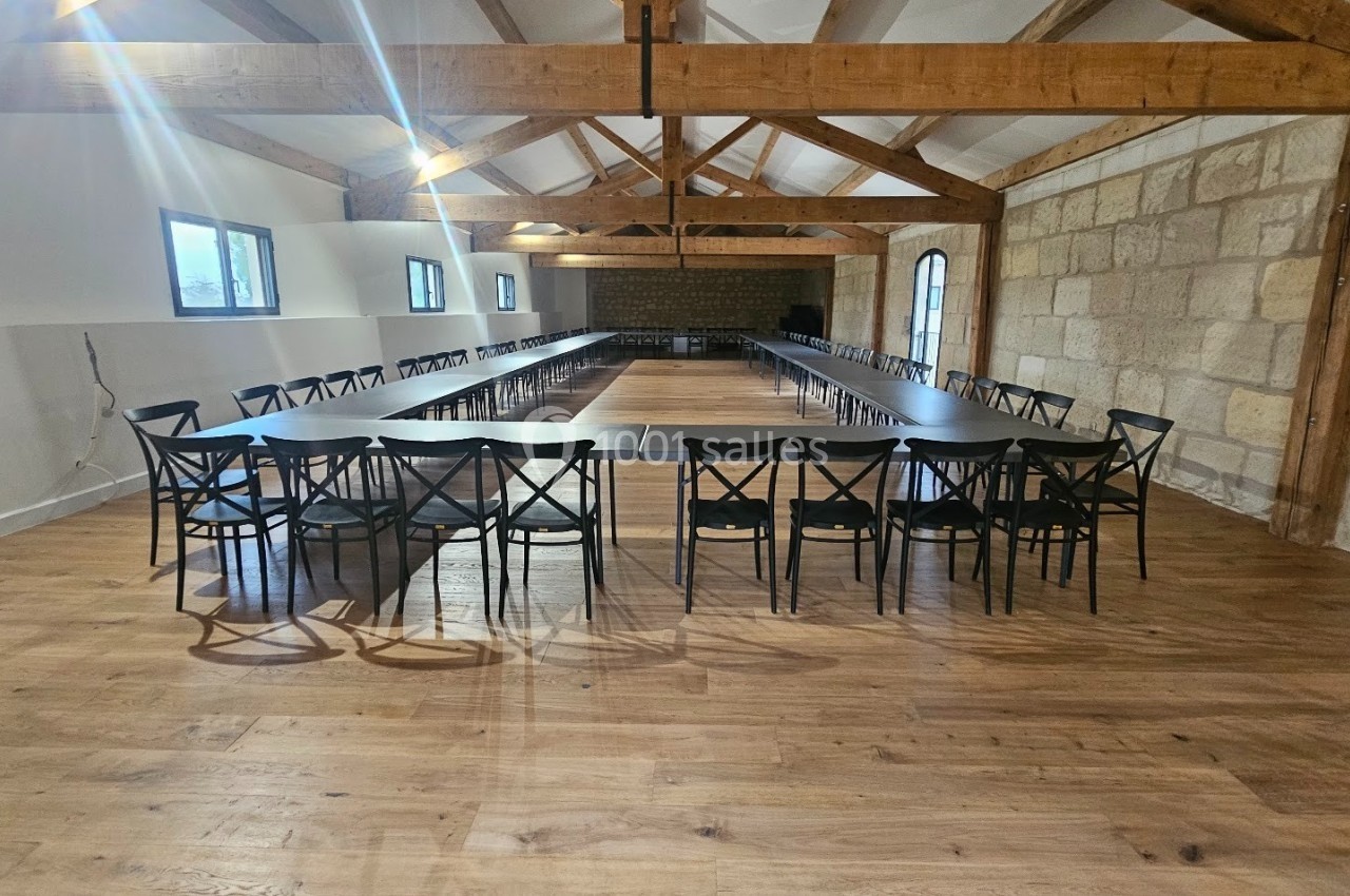 Salle spacieuse avec parquet en bois, poutres apparentes et tables disposées en U entourées de chaises noires.