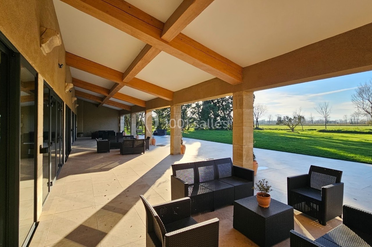 Terrasse couverte avec mobilier en rotin, donnant sur un grand jardin verdoyant sous un ciel dégagé.