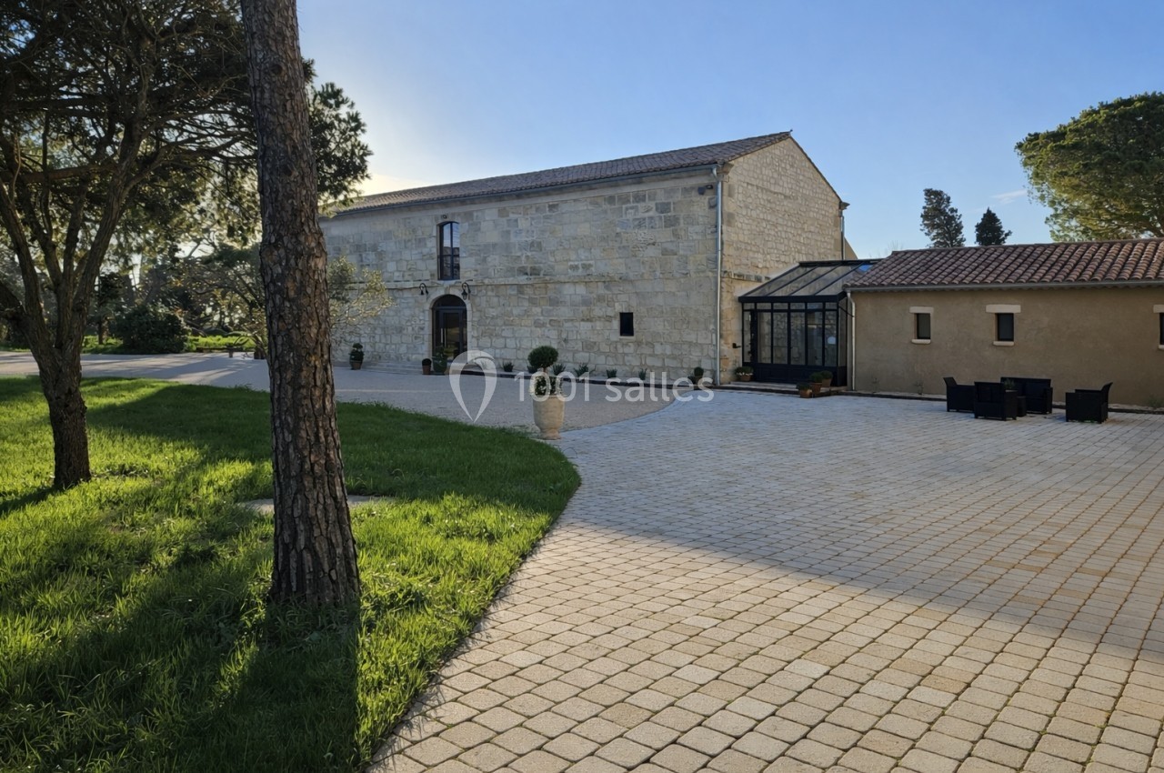 Bâtiment en pierre avec une véranda, entouré d'une cour pavée et d'arbres sous un ciel dégagé.