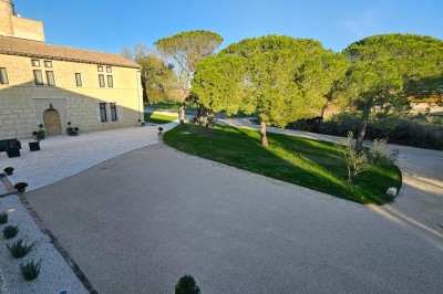 Cour extérieure d'une maison en pierre avec un arbre central, une véranda et des espaces aménagés sous un ciel dégagé.
