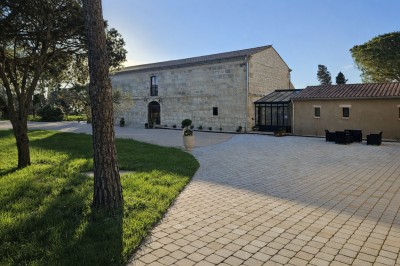 Cour extérieure d'une maison en pierre avec un arbre central, une véranda et des espaces aménagés sous un ciel dégagé.