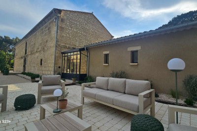 Cour extérieure d'une maison en pierre avec un arbre central, une véranda et des espaces aménagés sous un ciel dégagé.