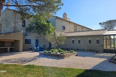 Cour extérieure d'une maison en pierre avec un arbre central, une véranda et des espaces aménagés sous un ciel dégagé.