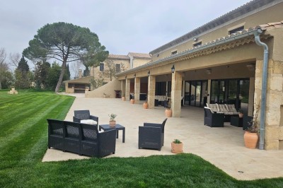 Cour extérieure d'une maison en pierre avec un arbre central, une véranda et des espaces aménagés sous un ciel dégagé.