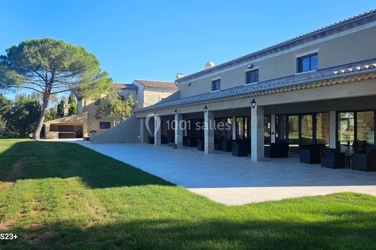 Grande maison moderne avec terrasse couverte, mobilier extérieur et jardin arboré sous un ciel dégagé.