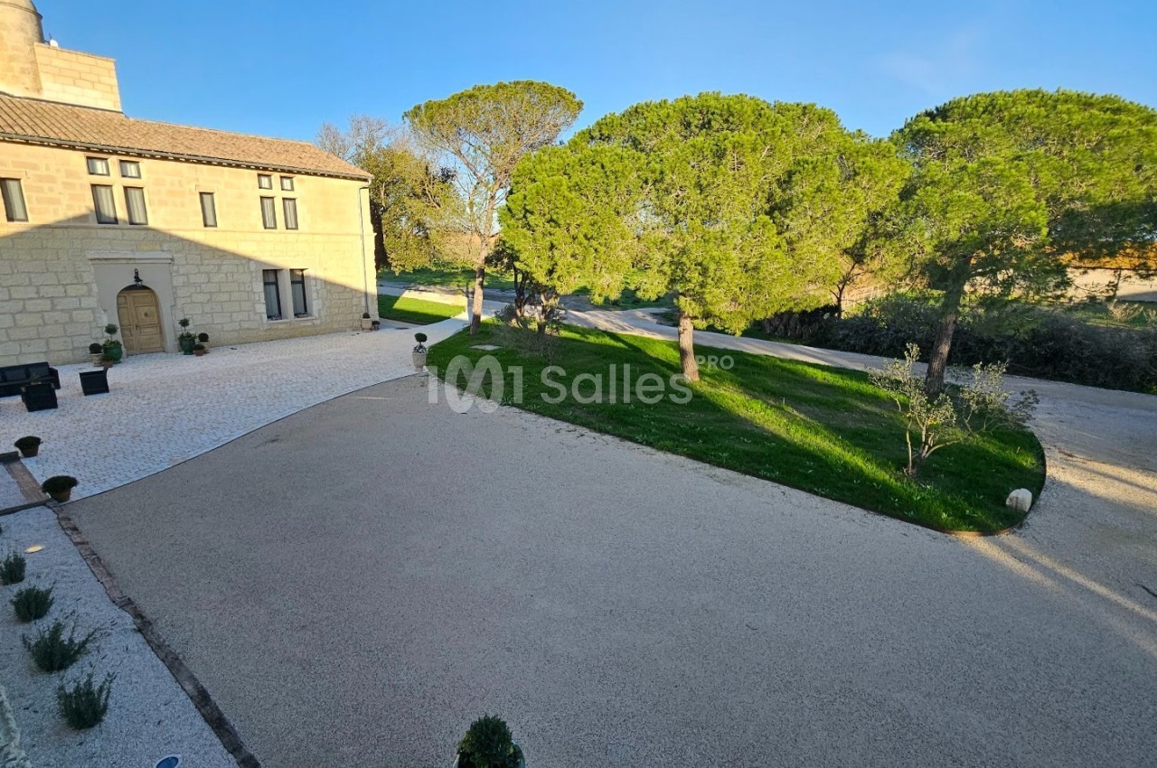 Allée gravillonnée bordée de pelouses et d'arbres devant une grande maison en pierre sous un ciel dégagé.