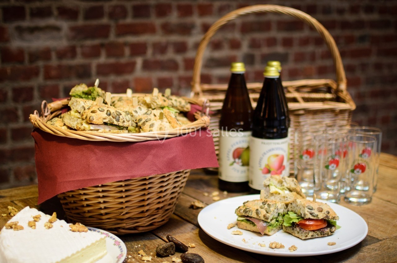 Panier de sandwichs garnis sur une table en bois avec bouteilles de jus, verres et fromage, devant un mur en briques.