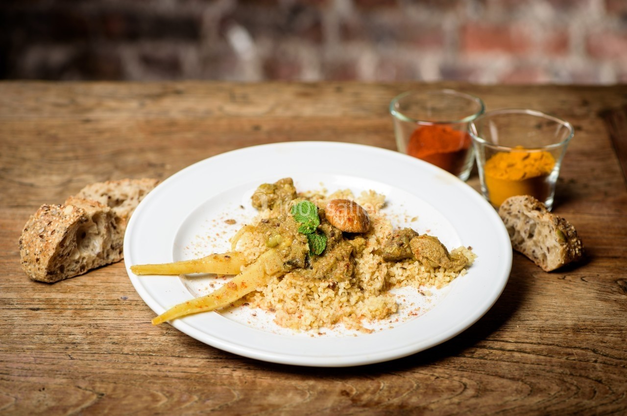Assiette de couscous avec légumes, viande et épices, servie sur une table en bois avec du pain et des épices en arrière-plan.