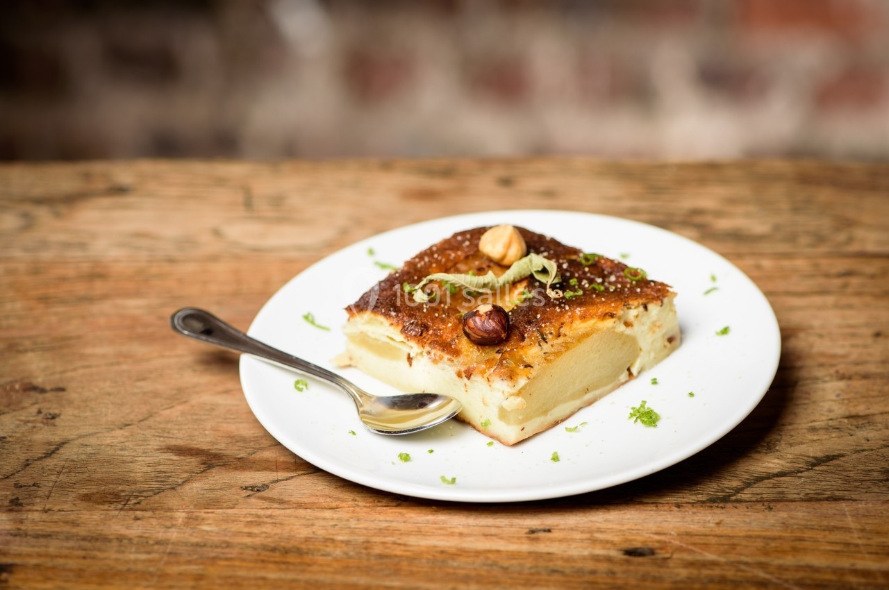 Part de gâteau doré sur une assiette blanche avec une cuillère, décorée de noisettes et de zestes verts.