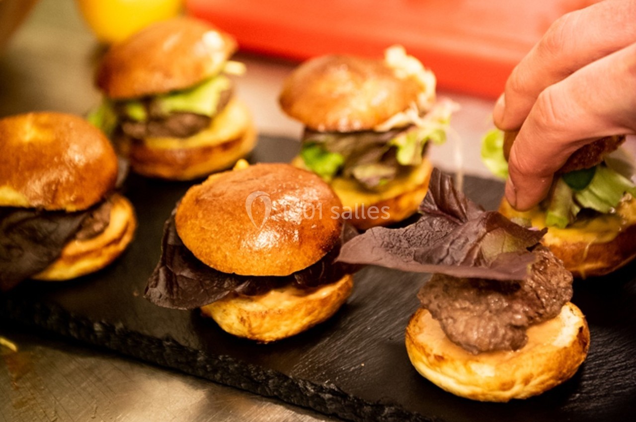 Mini burgers garnis de salade et viande, disposés sur une ardoise noire, avec une main ajustant une feuille de salade.