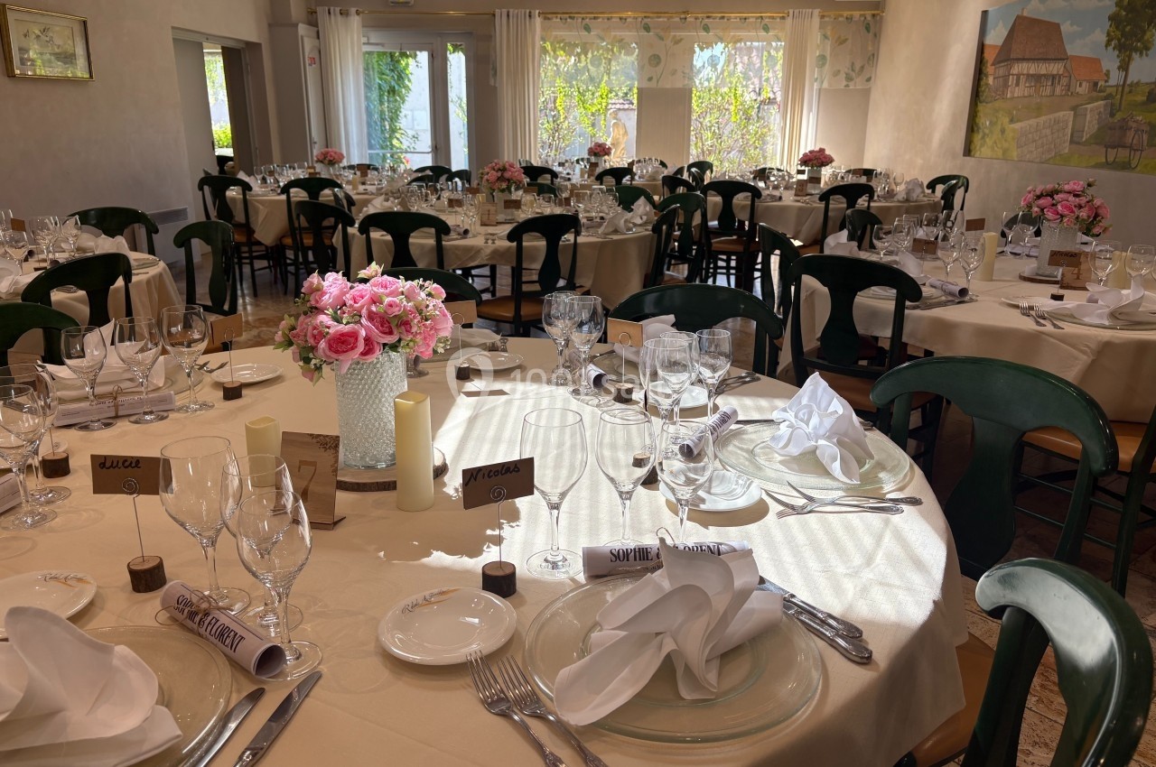 Salle de réception lumineuse avec tables rondes dressées, décorées de bouquets roses et serviettes pliées.